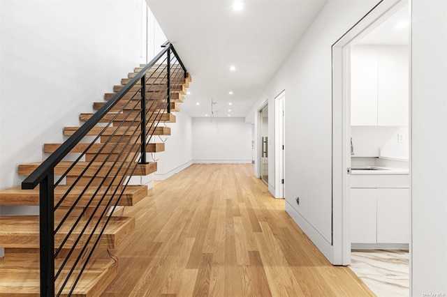 corridor featuring baseboards, light wood finished floors, recessed lighting, a sink, and stairs