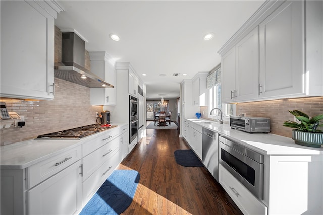 kitchen featuring a sink, wall chimney range hood, appliances with stainless steel finishes, light countertops, and dark wood-style flooring