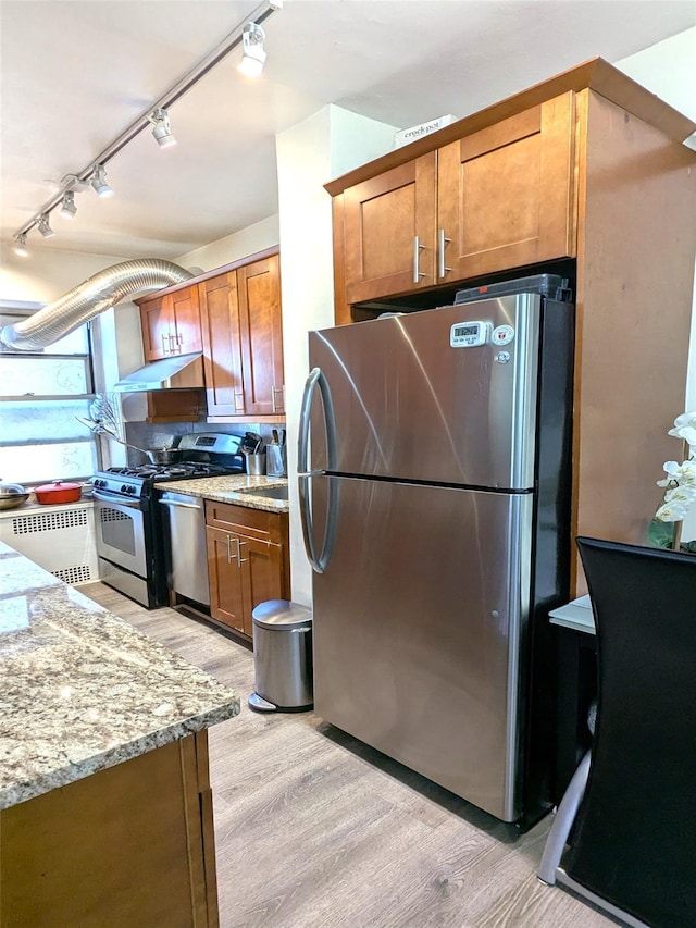 kitchen with under cabinet range hood, light stone counters, brown cabinets, light wood-style floors, and stainless steel appliances