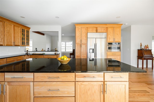 kitchen featuring light wood-type flooring, built in refrigerator, dark stone countertops, stainless steel oven, and glass insert cabinets