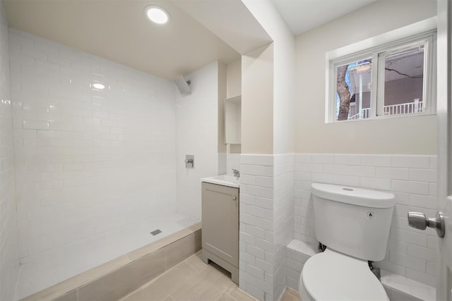 full bathroom with toilet, a tile shower, tile walls, wainscoting, and vanity