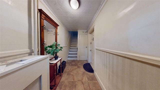 hallway featuring a wainscoted wall, a textured ceiling, light tile patterned flooring, crown molding, and stairs