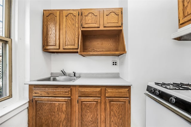 kitchen with white gas stove, under cabinet range hood, light countertops, brown cabinetry, and a sink