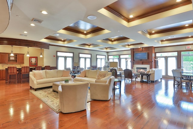 living area featuring wood finished floors, visible vents, coffered ceiling, ornamental molding, and a glass covered fireplace