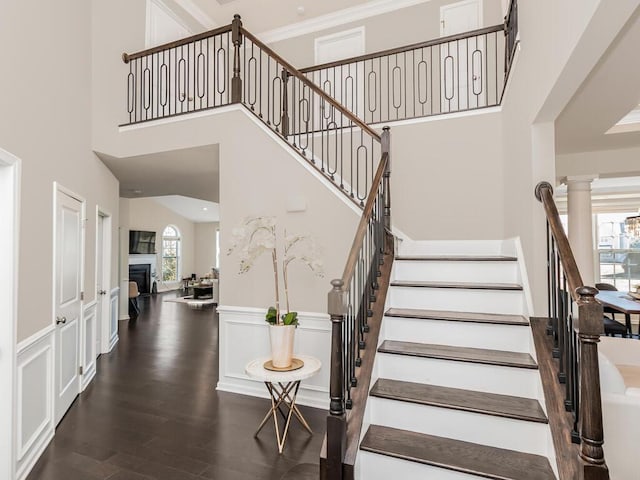 staircase featuring wood finished floors, ornate columns, a high ceiling, a fireplace, and crown molding