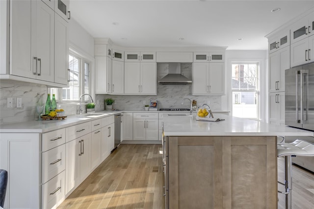 kitchen featuring a sink, wall chimney range hood, a kitchen breakfast bar, appliances with stainless steel finishes, and light wood finished floors