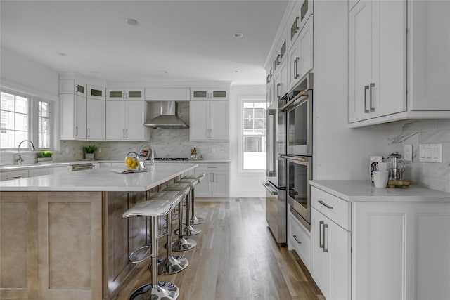 kitchen with a breakfast bar, a sink, glass insert cabinets, light wood-style floors, and wall chimney range hood