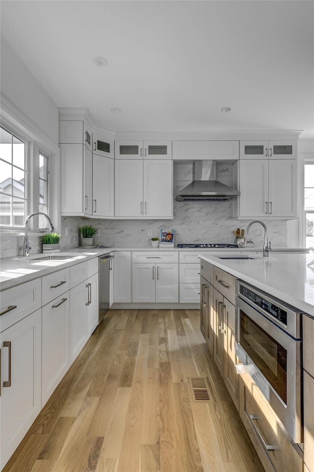 kitchen with a sink, wall chimney range hood, white cabinets, light countertops, and dishwasher