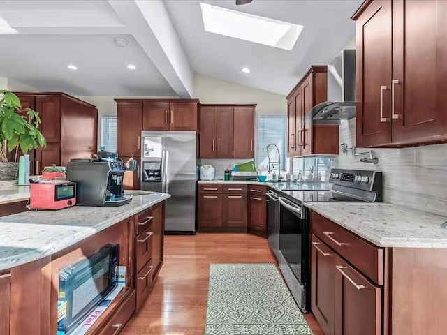 kitchen featuring lofted ceiling with skylight, stainless steel appliances, light wood-style floors, wall chimney range hood, and light stone countertops