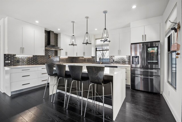 kitchen with wall chimney range hood, a center island, stainless steel fridge with ice dispenser, light countertops, and dark wood-style flooring