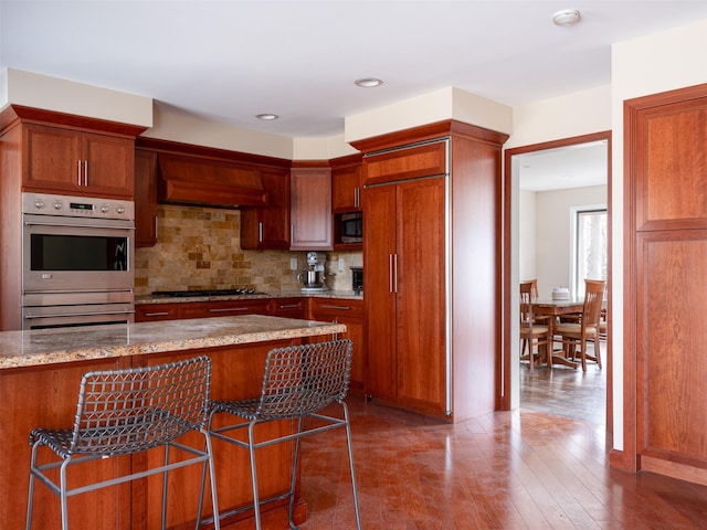 kitchen featuring premium range hood, light stone counters, backsplash, wood finished floors, and appliances with stainless steel finishes