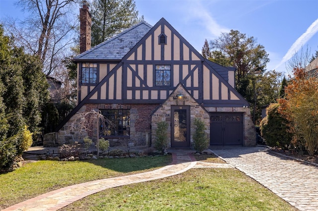 tudor-style house featuring driveway, a chimney, stucco siding, a front lawn, and stone siding