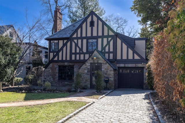 tudor home with stucco siding, decorative driveway, a front yard, a garage, and a chimney