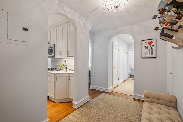 hall featuring electric panel, baseboards, arched walkways, and light wood finished floors