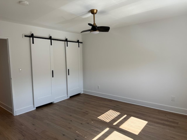 unfurnished bedroom featuring baseboards, a ceiling fan, a barn door, and dark wood-style flooring