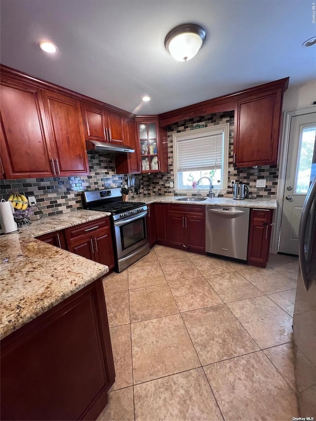 kitchen featuring light stone counters, a sink, under cabinet range hood, appliances with stainless steel finishes, and backsplash