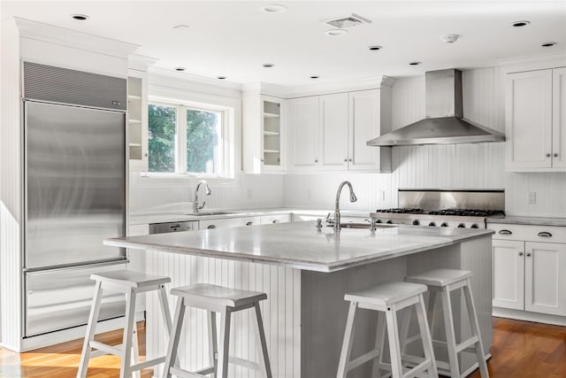 kitchen with visible vents, built in refrigerator, a sink, wall chimney range hood, and glass insert cabinets