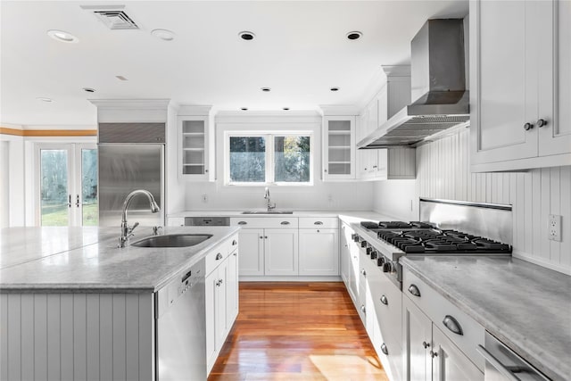 kitchen with a kitchen island with sink, a sink, appliances with stainless steel finishes, wall chimney range hood, and glass insert cabinets