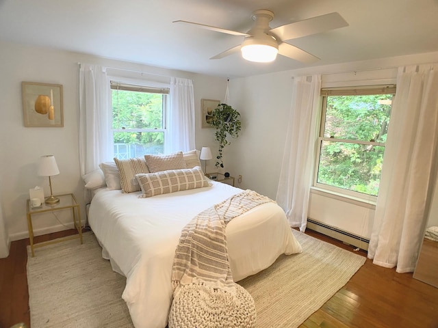 bedroom featuring baseboard heating, wood finished floors, ceiling fan, and multiple windows