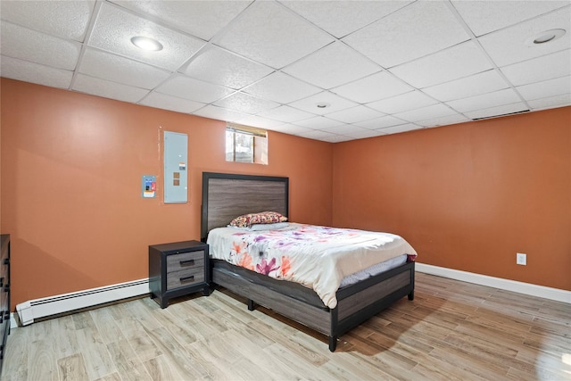 bedroom with electric panel, a drop ceiling, a baseboard heating unit, light wood-style floors, and baseboards