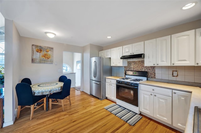 kitchen with white gas stove, under cabinet range hood, white cabinetry, freestanding refrigerator, and decorative backsplash