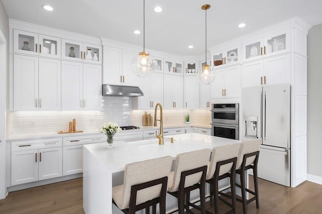 kitchen featuring dark wood-type flooring, a center island with sink, under cabinet range hood, stainless steel double oven, and white fridge with ice dispenser