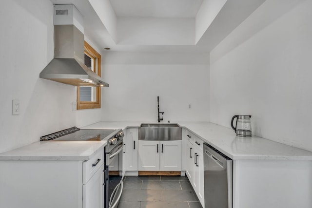 kitchen with light stone counters, white cabinets, stainless steel appliances, wall chimney exhaust hood, and a sink