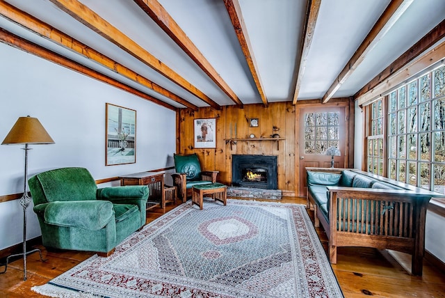 living area featuring beam ceiling, wooden walls, a warm lit fireplace, and wood-type flooring