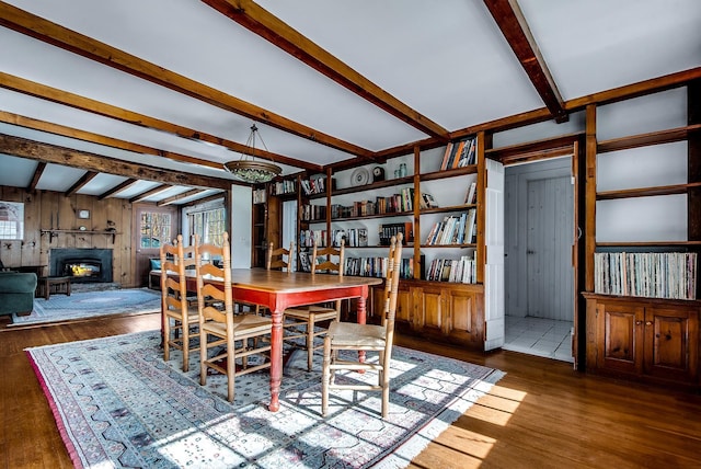 dining space with beam ceiling, wooden walls, a glass covered fireplace, and wood finished floors