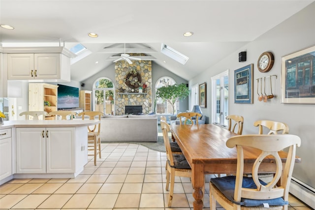 dining area with vaulted ceiling with skylight, a stone fireplace, light tile patterned floors, a baseboard radiator, and ceiling fan