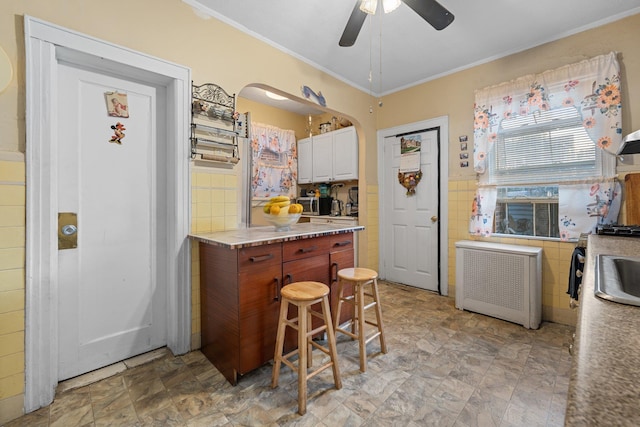 kitchen with ornamental molding, white cabinetry, a ceiling fan, radiator, and tile walls