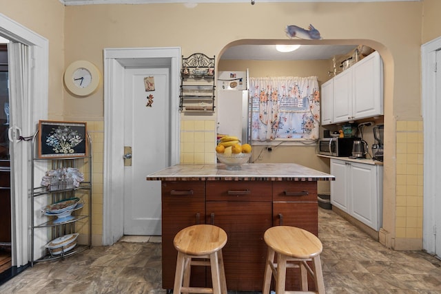 kitchen featuring white cabinets, arched walkways, light countertops, tile walls, and stainless steel microwave