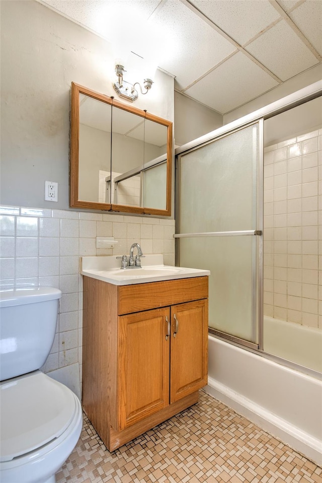 bathroom with toilet, a drop ceiling, tile walls, bath / shower combo with glass door, and vanity