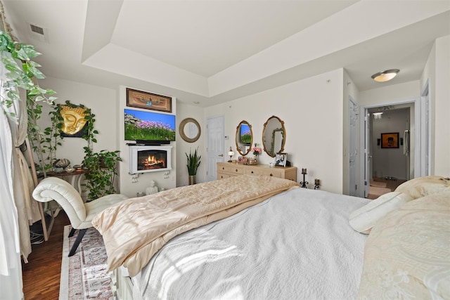 bedroom featuring visible vents, a warm lit fireplace, a tray ceiling, and wood finished floors