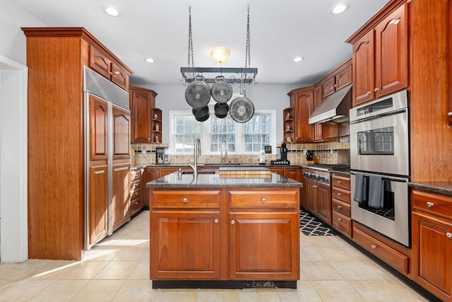 kitchen with under cabinet range hood, dark stone countertops, appliances with stainless steel finishes, and an island with sink