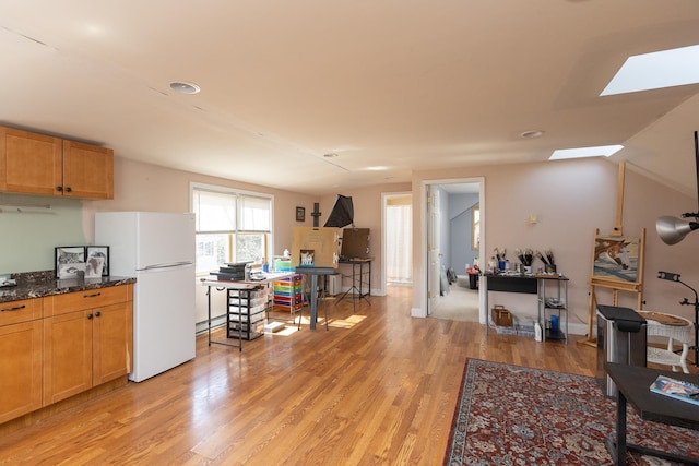 kitchen with light wood-type flooring, freestanding refrigerator, dark stone counters, vaulted ceiling with skylight, and baseboards