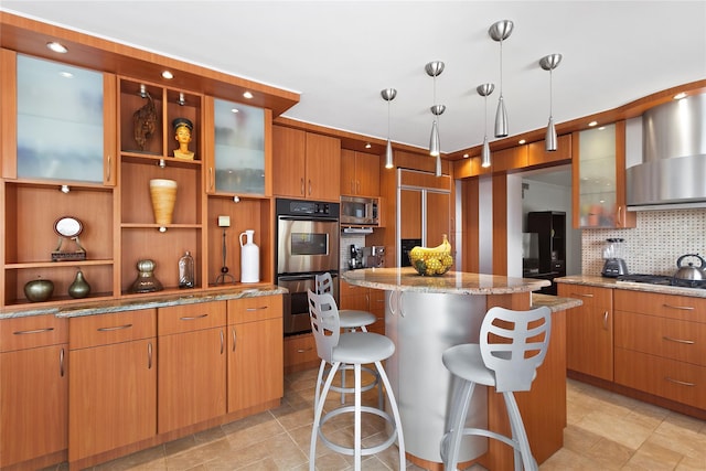 kitchen featuring a kitchen breakfast bar, a center island, stainless steel appliances, wall chimney range hood, and light stone countertops