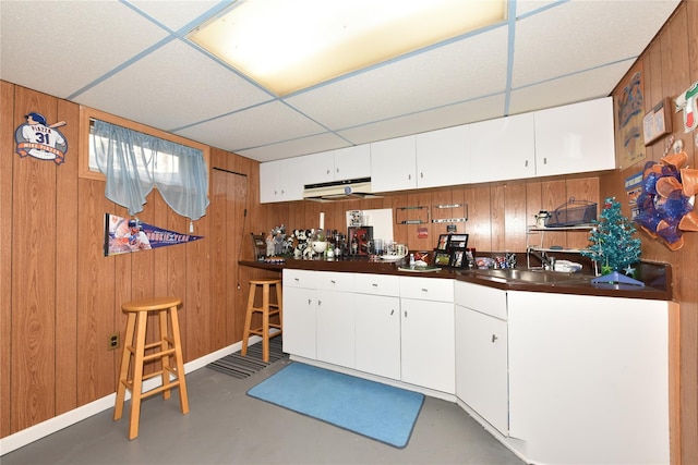 kitchen with a sink, dark countertops, wood walls, a paneled ceiling, and concrete flooring