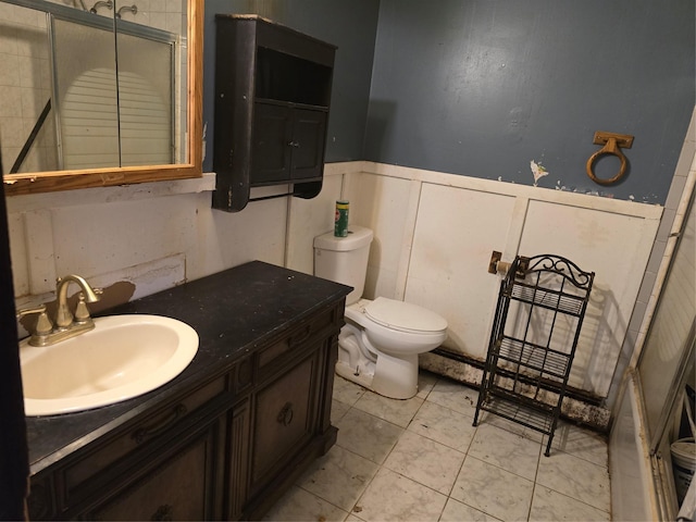 full bath with a shower with shower door, a wainscoted wall, toilet, tile patterned flooring, and vanity