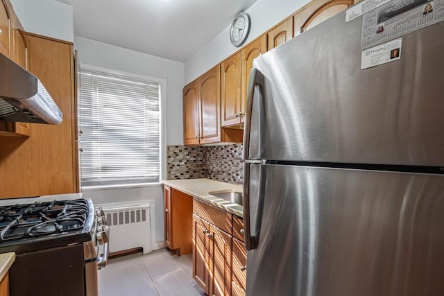 kitchen featuring brown cabinets, under cabinet range hood, tasteful backsplash, radiator, and appliances with stainless steel finishes