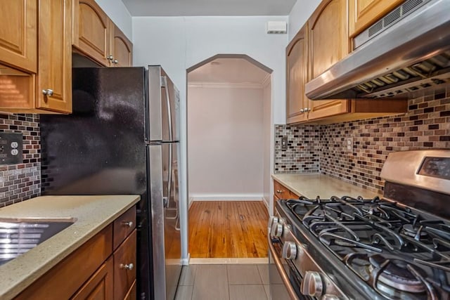 kitchen featuring under cabinet range hood, light stone counters, arched walkways, appliances with stainless steel finishes, and decorative backsplash