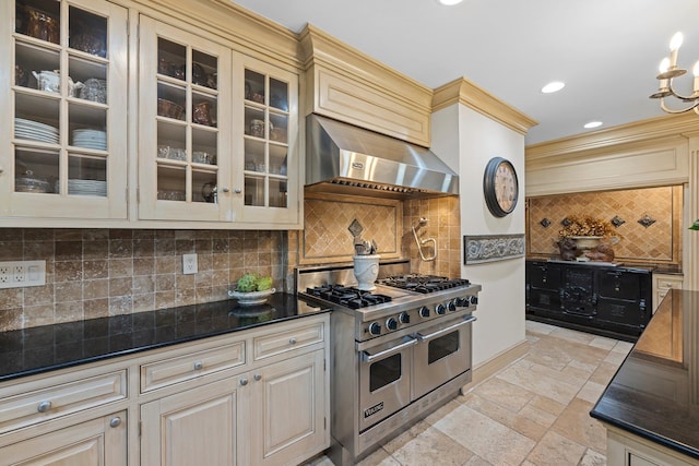 kitchen with glass insert cabinets, wall chimney range hood, double oven range, stone tile floors, and cream cabinets