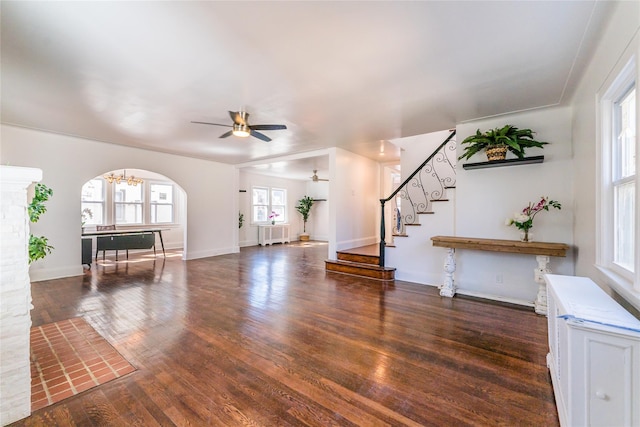 unfurnished living room featuring radiator, wood finished floors, arched walkways, stairs, and ceiling fan with notable chandelier