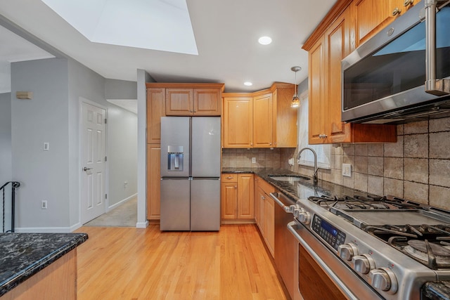 kitchen featuring light wood finished floors, dark stone counters, a skylight, a sink, and appliances with stainless steel finishes