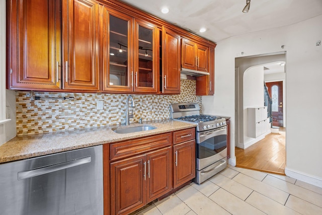 kitchen with a sink, arched walkways, appliances with stainless steel finishes, glass insert cabinets, and light stone countertops