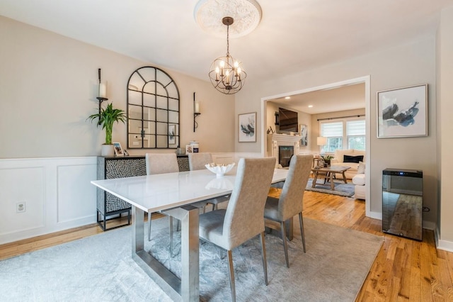 dining room with a wainscoted wall, light wood finished floors, a notable chandelier, a glass covered fireplace, and a decorative wall