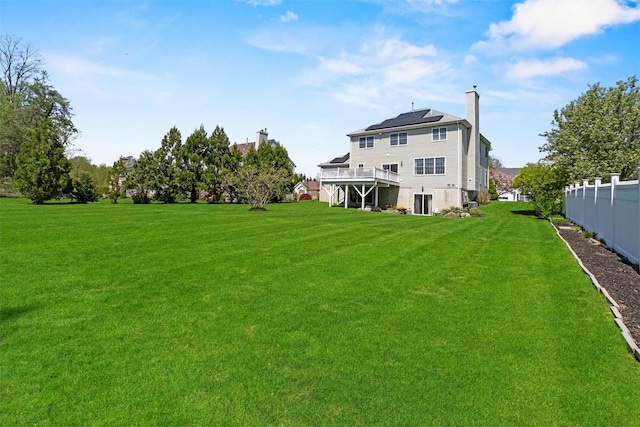 rear view of property featuring roof mounted solar panels, a chimney, fence, a lawn, and a deck