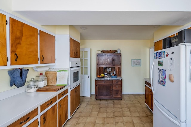 kitchen featuring light countertops, baseboards, brown cabinetry, and white appliances