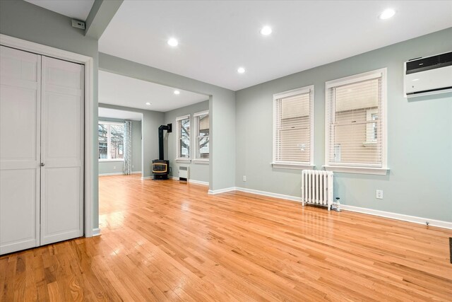 89-21 Aubrey Ave, Glendale, NY 11385 - unfurnished living room featuring radiator heating unit, a wood stove, light wood-style floors, a wall unit AC, and baseboards