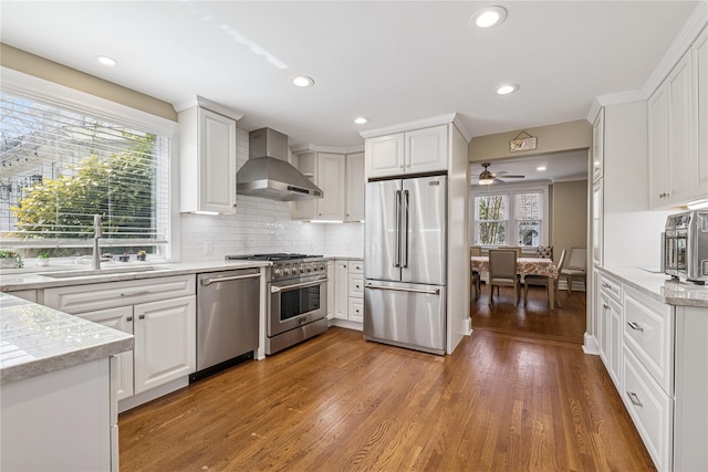 kitchen with wall chimney range hood, a sink, ceiling fan, high end appliances, and white cabinets
